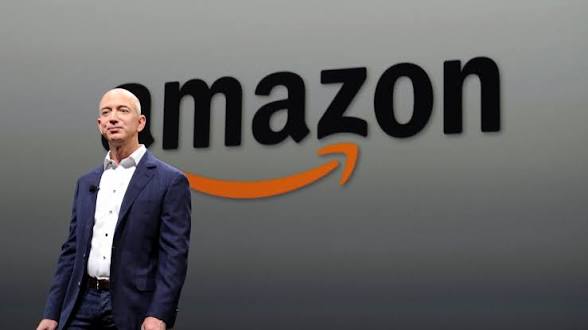 Jeff Bezos in a navy suit standing in front of the Amazon logo